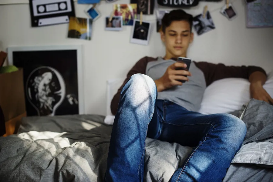 Tenåring sitter avslappet på sengen sin og scroller på mobilen.