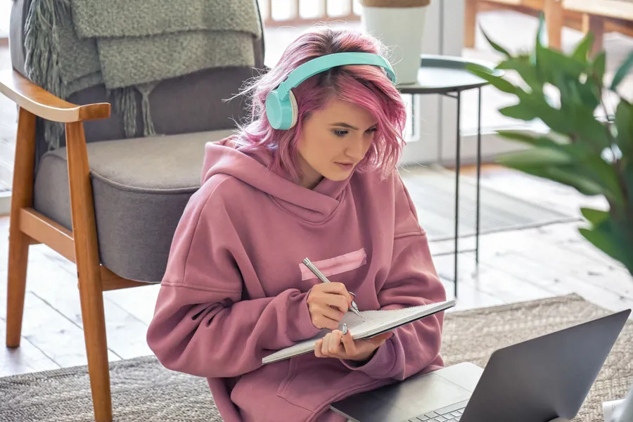 Ung jente sitter med hodetelefoner og notatblokk hjemme i stuen sin. Har laptop foran seg og tar notater fra skjermen.
