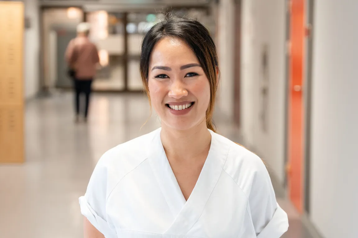 Sykepleier kledd i hvit uniform står smilende i en korridor på sykehuset.