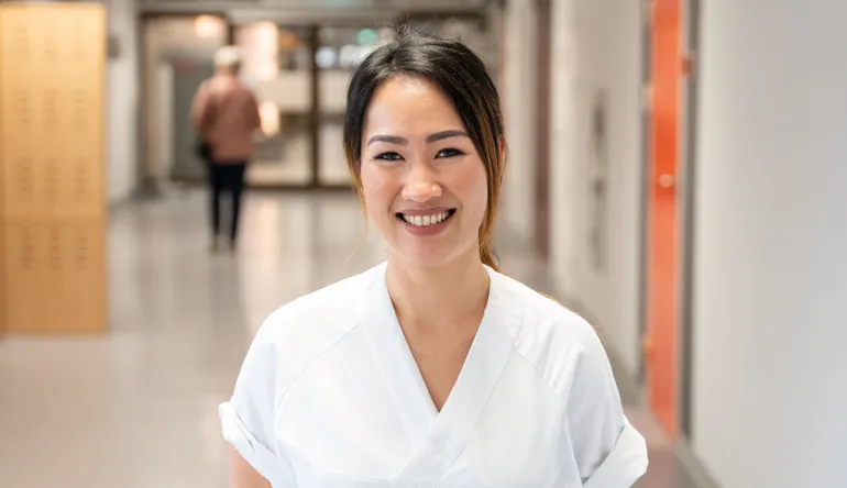 Sykepleier kledd i hvit uniform står smilende i en korridor på sykehuset.