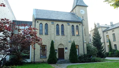 Kirkebygg fra 1881 oppført i gul murstein. Bygget er omgitt av gress og trær. Foto.