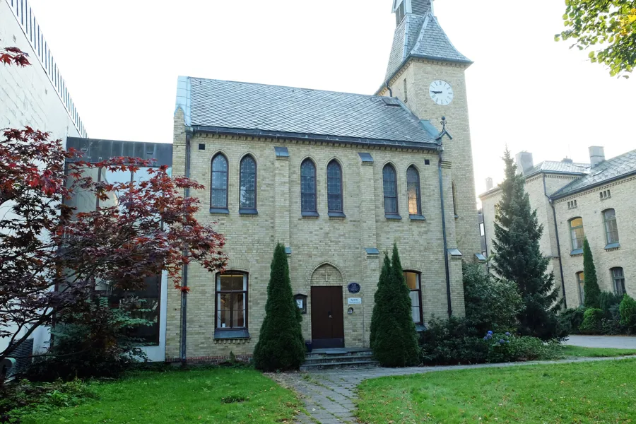 Kirkebygg fra 1881 oppført i gul murstein. Bygget er omgitt av gress og trær. Foto.