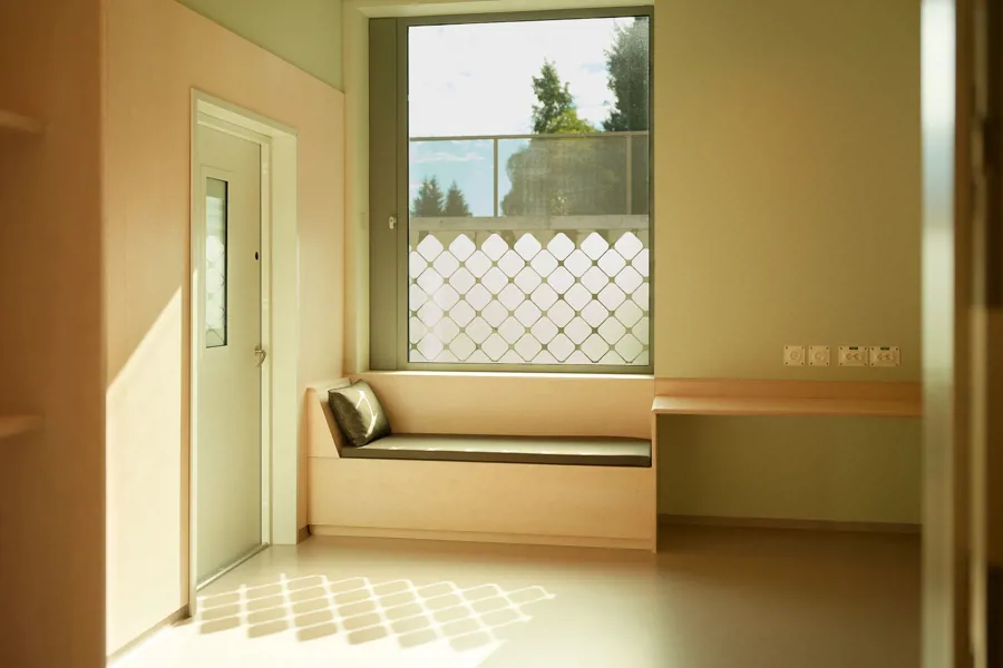 Pasientrom med stort vindu, daybed og skrivebord. Sollyset reflekteres i rommet. Foto.